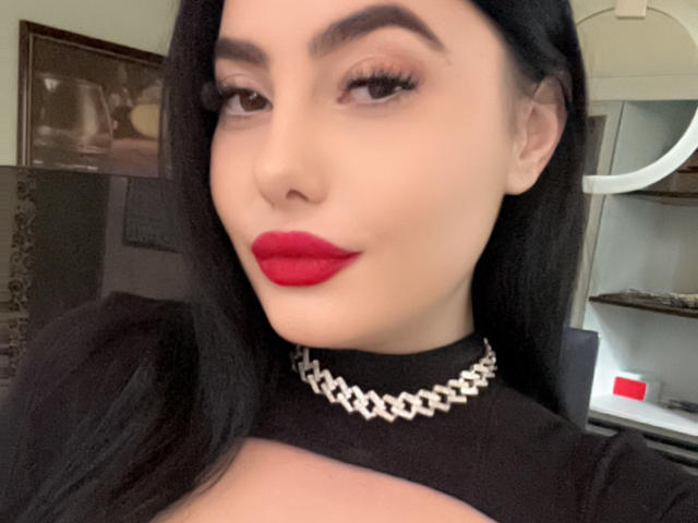 MelissaGlow has pale skin, black hair, red lipstick, and wears a silver chain necklace with a black top. Her boobs are visible in the shot.