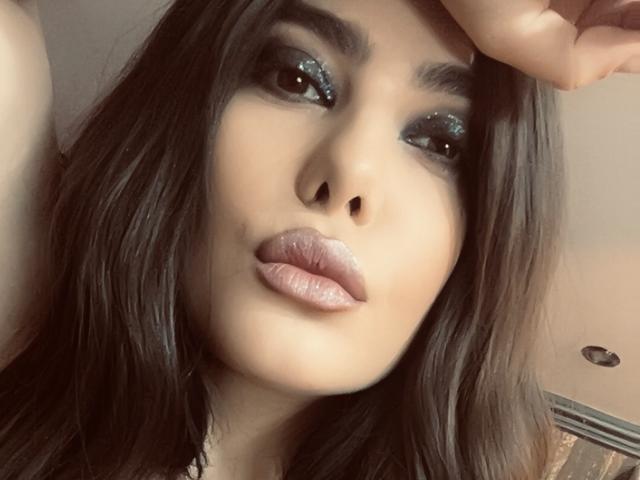 MelissaGlow's face with dark brown hair, glittery blue eyeshadow, and full pink lips. She's looking at the camera with a slight pout, hand resting on her head.