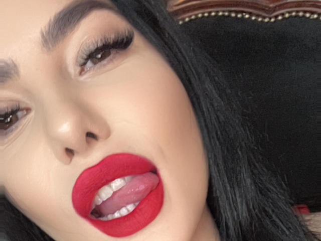 MelissaGlow's face with red lipstick, black hair, and tongue slightly out, showing teeth. She's got long eyelashes and a slightly open mouth, looking sexy and seductive.