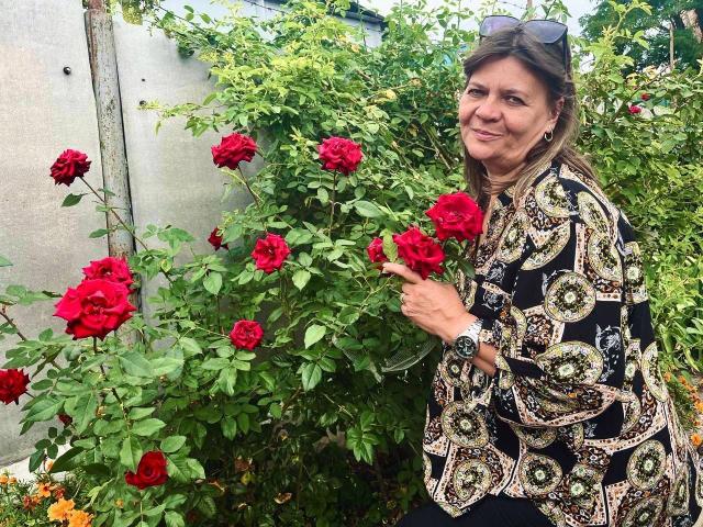 LolaSun with red roses, wearing a black patterned blouse, smiling, pointing at flowers, wearing a watch, sunglasses on head, in a garden.