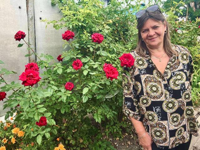 LolaSun with medium tits, smiling, wearing a patterned black and green blouse, standing beside a red rose bush, wearing sunglasses on head, in a sunny garden.