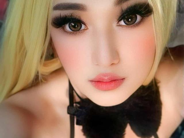 QuuenBeeTs has blonde hair, heavy black eyeliner, and pink lipstick. She's looking directly at the camera with wide brown eyes. She's wearing a black furry collar.