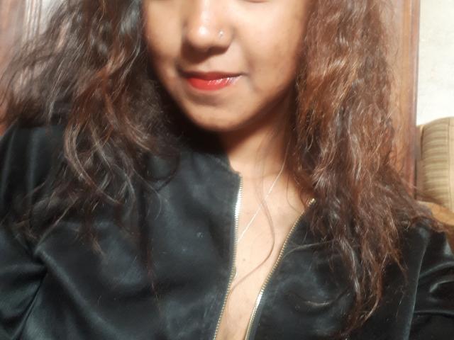 JudeXi with wavy brown hair, red lipstick, black zip-up shirt, nose piercing, and a subtle smile, showing a little cleavage. Her tits are small and her pussy is not visible.