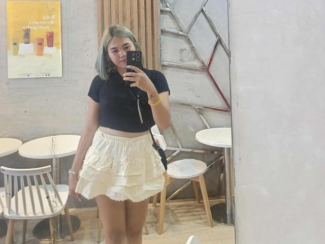 AsianHotCock69 with pale green hair, black crop top, and white ruffled skirt, taking a mirror selfie, holding a phone with a yellow bracelet.