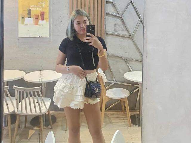 AsianHotCock69 with gray hair, black crop top, white ruffled skirt, black purse, holding phone, standing in cafe, mirror selfie, showing ass, with bracelet, looking at camera.