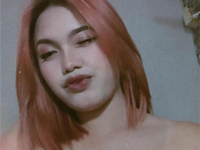 AsianHotCock69 with pink hair, light skin, and pouty lips, slightly squinting eyes, topless, showing bare shoulders, soft light, subtle shadow, subtle smile, slightly blurred photo.