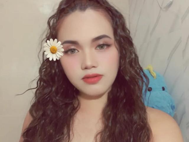 AsianHotCock69 has curly brown hair, pale skin, and is wearing a daisy in her hair with pink lipstick and subtle makeup. She's looking directly at the camera.