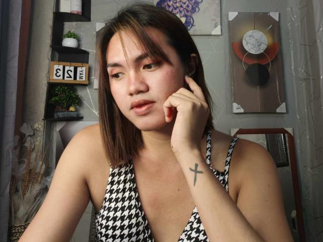 AsianHotCock69 with short brown hair, wearing a black-and-white houndstooth dress, has a tattooed cross on her right wrist, resting her chin on her hand, looking slightly bored.