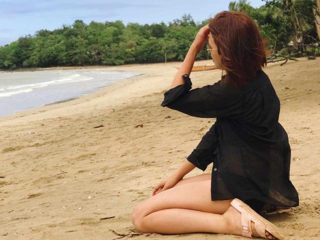 AsianHotCock69, sitting on sandy beach, wearing black shirt, no pants, pink sandals, brown hair, hand in hair, looking at ocean, legs crossed, soft sand, green trees in background.