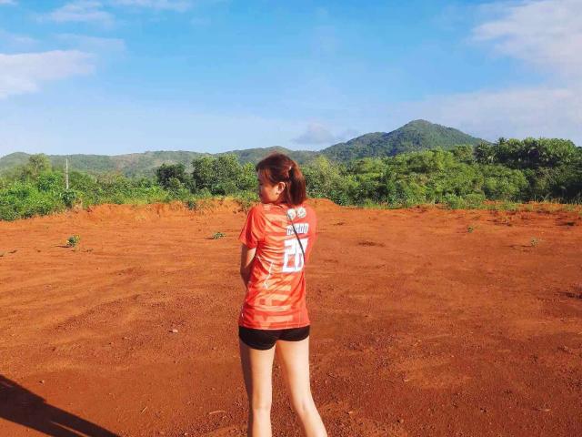 AsianHotCock69, standing with back to camera, wearing orange shirt and black shorts, on red dirt field, with green hills and blue sky in background. Red shirt has white text.