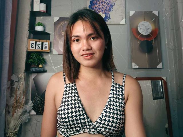 AsianHotCock69 with medium tits, wearing a black-and-white houndstooth bra, has shoulder-length brown hair, and a slight smile, standing in a room with decorative art.