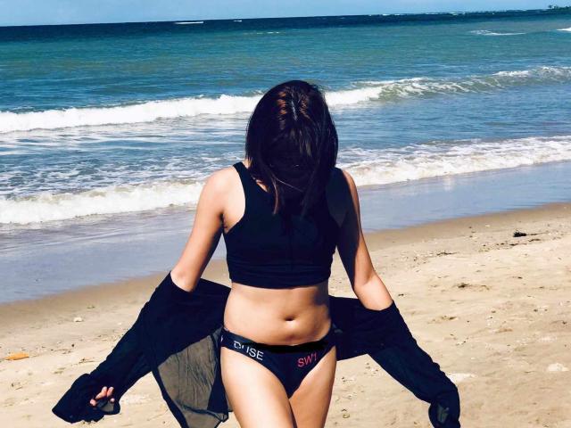 AsianHotCock69 with black sports bra and black "SLIM" panties, holding a black shirt, standing on sandy beach, facing ocean, mid-day sunlight, brunette, medium build.