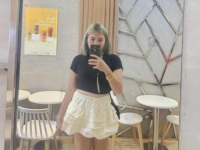 AsianHotCock69 with green-dyed bob, black crop top, and white ruffled skirt, holding phone, mirror selfie, visible tattoo on left forearm, wearing yellow bracelet.