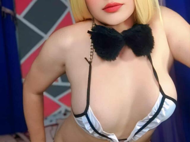 QuuenBeeTs with blonde hair, red lipstick, wearing a black furry bowtie, and a white, black-trimmed, barely-there bikini, showing her big, round tits.