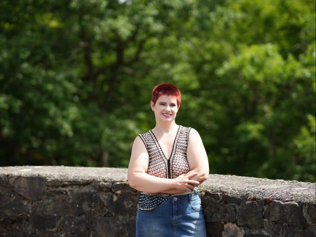 3LochStute-hot with red hair, smiling, wearing a sleeveless, patterned top and blue jeans, stands in front of a stone wall, arms crossed, showing her tits.