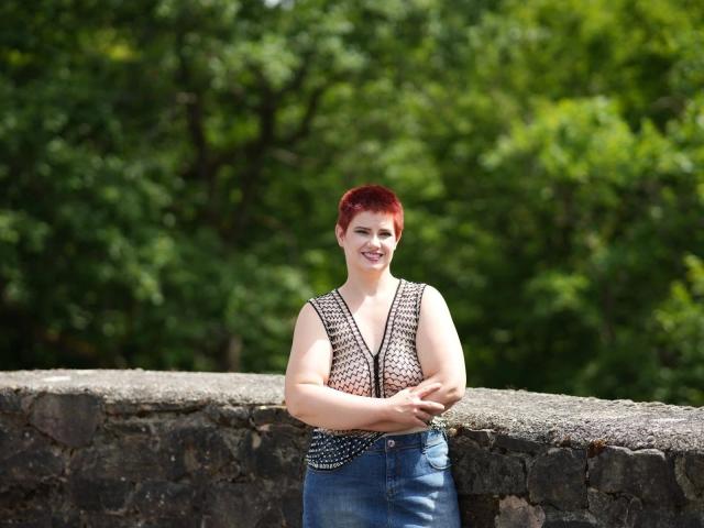 3LochStute-hot with red hair, sleeveless black and white patterned top, and blue jeans, standing in front of a stone wall, smiling, arms crossed.