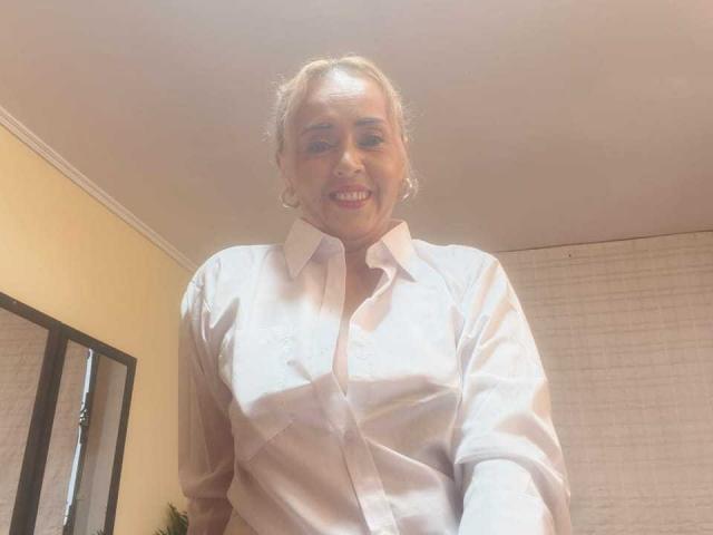 MaylinYung with blonde hair, wearing a white button-up shirt, smiling, showing small tits, and wearing hoop earrings, in a brightly lit room.