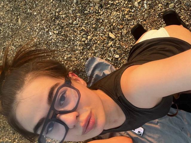 EvaStoneX with glasses, lying on a pebbled beach, black top, white panties, black socks, brown hair in a bun, looking at the camera, butt slightly raised.