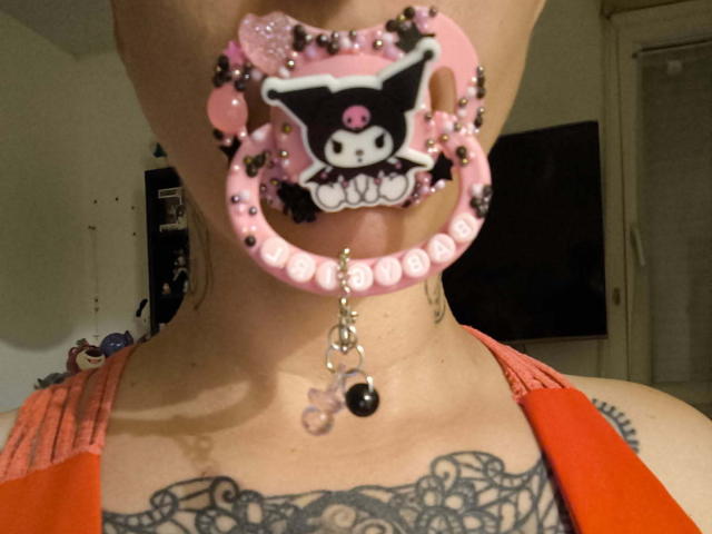 MissLilis has a pink Hello Kitty lip ring with beads, a dangling chain, and tattoos on her neck, wearing an orange top. Her tongue is visible.