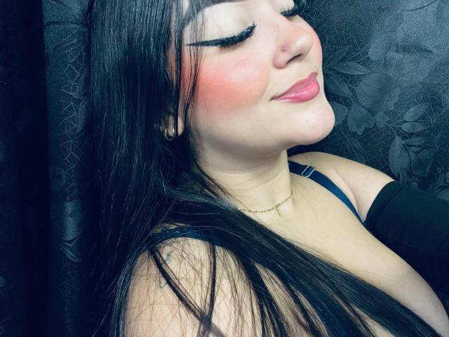 AnaShalimarr, com longos cabelos pretos, blush rosa e lábios rosados brilhantes, sorrindo com os olhos fechados, vestindo alças pretas, um piercing no nariz e um colar de ouro, com um fundo escuro estampado com flores..