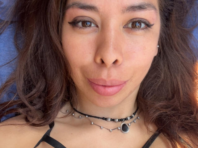AyeBeauty has brown eyes, pink lips, and a subtle smile. She wears a black necklace with silver beads and a small black gem. Her brown hair is wavy.