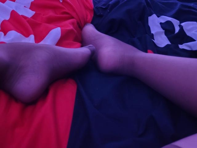ElylaFrenchy's soles are touching, her feet crossed on a red and black bed. Her toes are slightly curled, and the image has a purple-blue tint.
