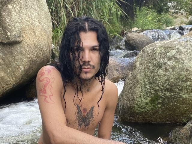 OshunRios with long wet black hair, tattoos on shoulder and chest, sitting in a rocky stream, looking at camera, shirtless, with a piercing on lower lip.