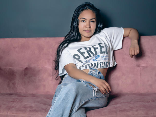 NatallyCooper with long black hair, wearing a white crop top and blue jeans, sitting on a pink couch, arm resting on the back, looking at the camera, slight smile.