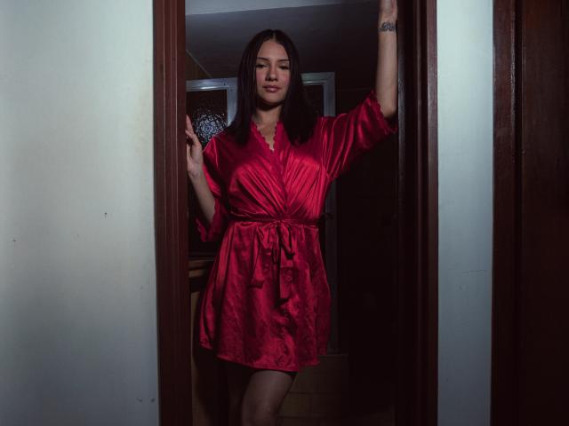 LizzJoy stands in a doorway wearing a red satin dress, holding a blue fan, with one arm raised, showing a tattoo on her wrist. Her tits are visible.