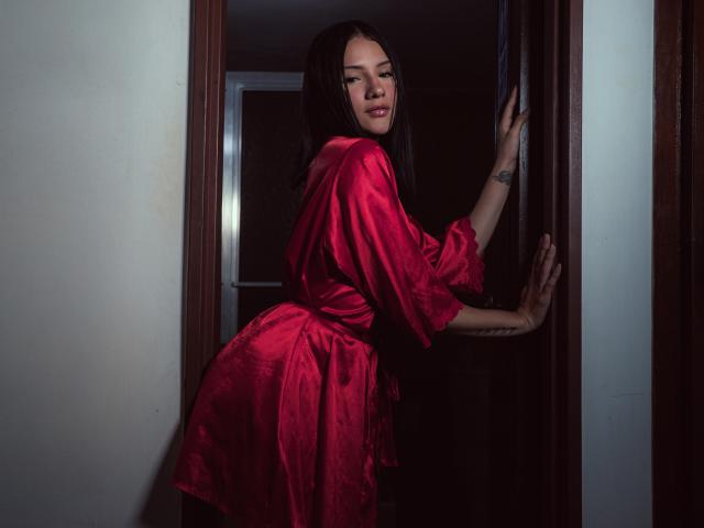 LizzeJoy in a shiny red satin dress, leaning against a dark wooden door, looking over her shoulder with a slight smile, showing her tattooed arm and red lips.