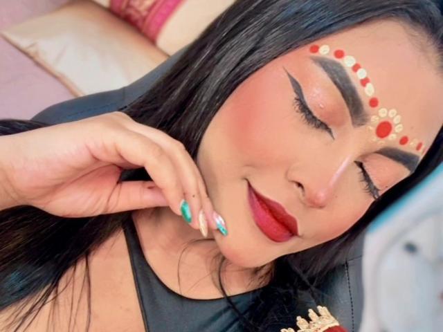 LakshmIrani has dark hair, closed eyes, red lipstick, and bold eyeliner. She's wearing a red and gold forehead ornament with red dots, and has teal and gold nails.