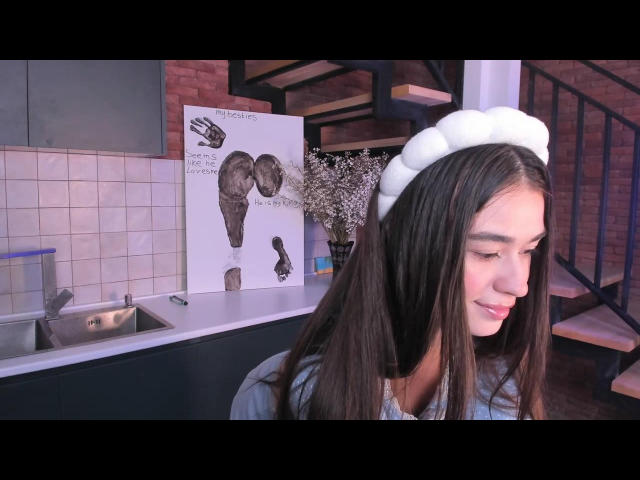 EvaStoneX with long brown hair, white fluffy headband, and subtle smile, standing in a kitchen, looking down, wearing a gray shirt, next to a sink and artwork.