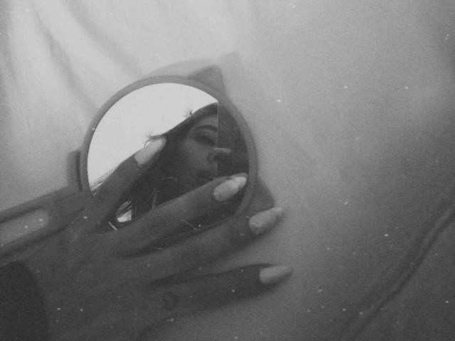 CattsyCathy's reflection in a round mirror, showing her face with long nails, in a grayscale, slightly grainy photo. Her hand is holding the mirror.