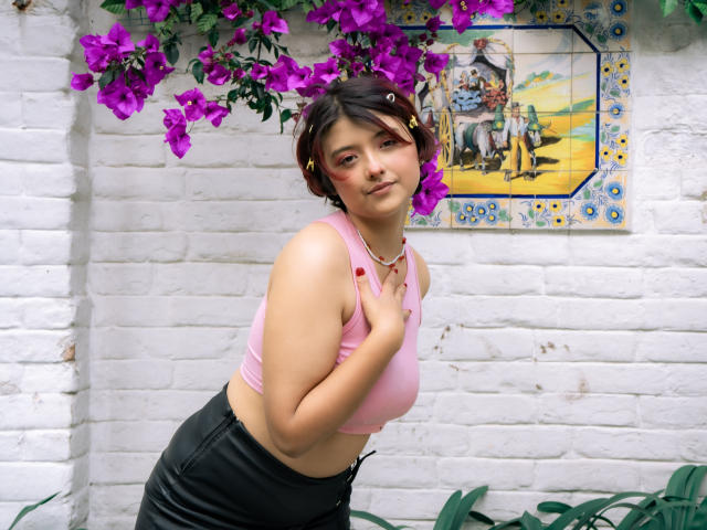 MisakiWong with short brown hair, pink crop top, showing small tits, black leather pants, standing in front of white brick wall, purple bougainvillea, and colorful tile.