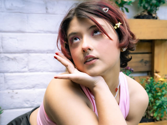 MisakiWong with short red-brown hair, pink lipstick, and a pink top, resting her cheek on her hand, looking up, showing a cute nose ring and a small flower hairpin.