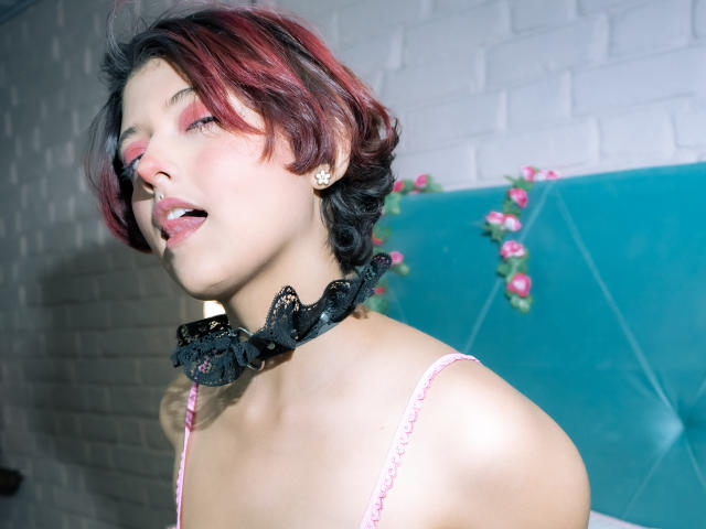 MisakiWong with red and black hair, pink makeup, black floral choker, and pink spaghetti strap top, tongue slightly out, looking sexy, and showing a tattoo on her neck.