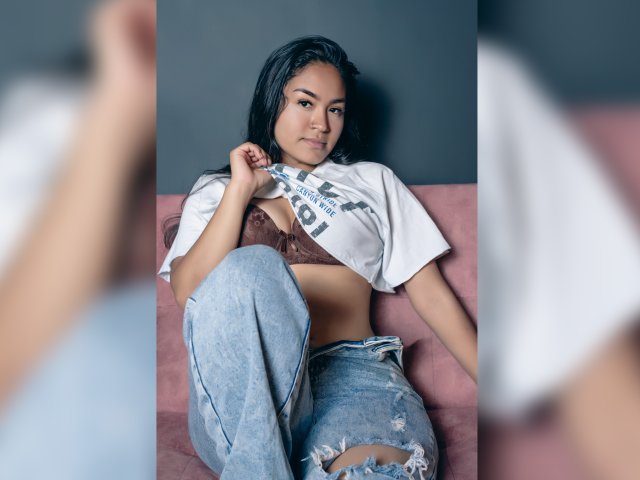 NatallyCooper with dark hair, brown bra, white shirt, and ripped jeans, sitting on pink couch, lifting shirt to show tits, looking at camera with a slight smile.