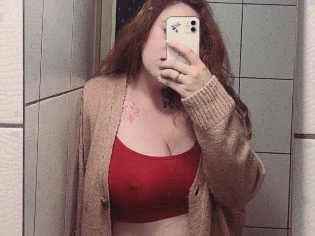 FortuneCarmen with red bra, showing her tits, wearing a beige cardigan, holding a phone in a mirror selfie, long red hair, tattoo on right chest.