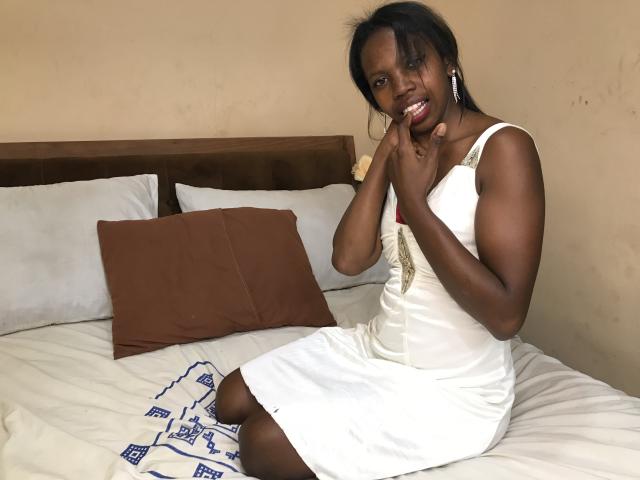 ElianeJolie, dark-skinned woman, white dress, sitting on bed, smiling, finger in mouth, brown pillows, white bedsheet, blue patterned blanket, brown headboard, black hair, medium-sized tits.