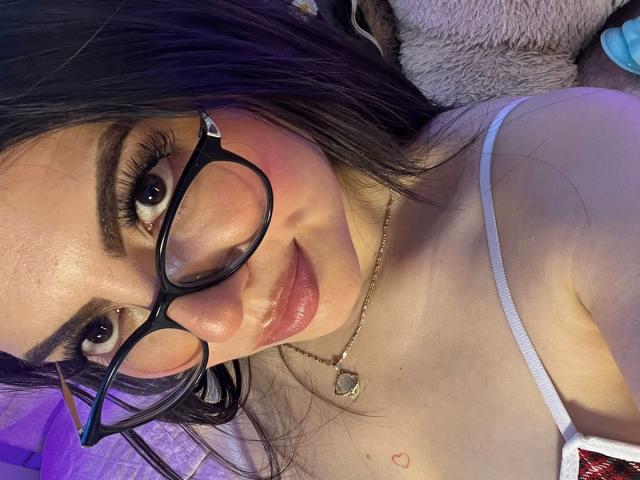 DorothyLane, usando óculos, com blush rosa e lábios brilhantes, vestindo uma alça branca, um colar dourado e uma tatuagem de coração perto do seio. Ela está olhando para a câmera com um leve sorriso..