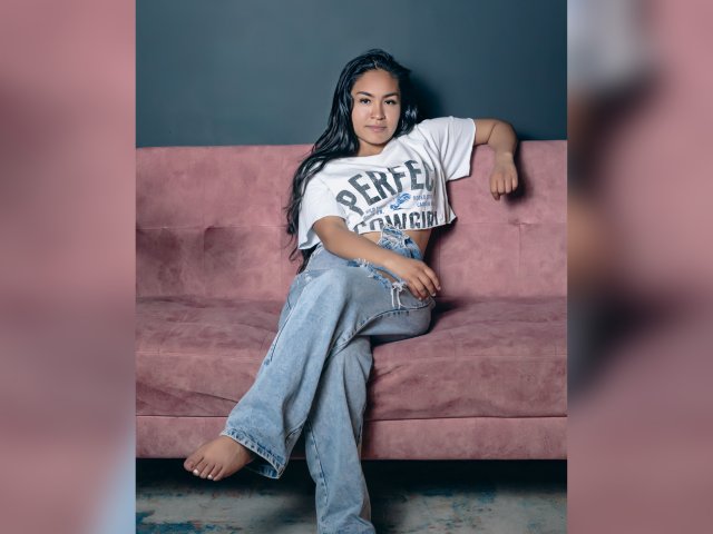 NatallyCooper with long black hair, wearing a white "PERFECT" t-shirt and loose blue jeans, leaning on a pink couch, barefoot, looking at the camera, relaxed, confident.