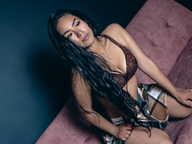 Natally Cooper met lang, zwart, nat haar, een bruine kanten bh, een geruite rok, zittend op een roze bank, kijkend naar de camera, donkere achtergrond, lichte glimlach, handen op de knieën, subtiele make-up..