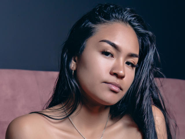 NatallyCooper has smooth brown skin, long black hair, and is looking slightly down with a serious expression. She's topless with small, perky tits and a silver necklace.