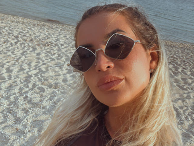 Lellie with blonde hair, wearing dark sunglasses, has a pouty face and a tan skin tone. She's on a sandy beach with a blue water background.