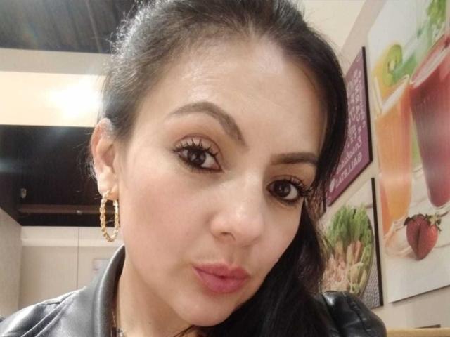 JakyKidman has dark, wavy hair, brown eyes, and is pouting with pink lips. She wears gold hoop earrings and a black leather jacket. Her makeup is subtle and sexy.