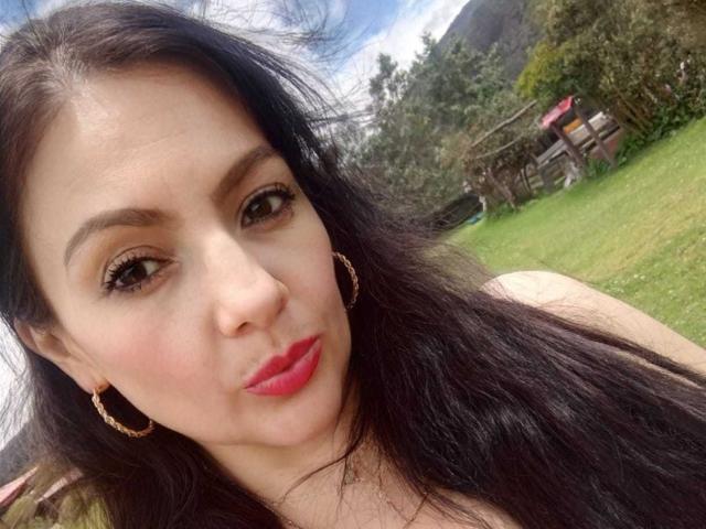 JakyKidman has long black hair, brown eyes, red lipstick, and gold hoop earrings, with a slightly pouty face and a small nose. She's outdoors on a sunny day.