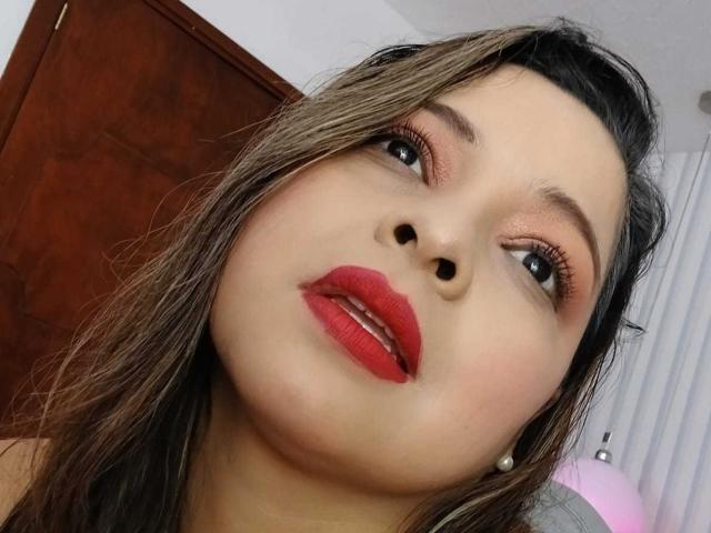 IdyllaJhones has red lipstick, full lips, and brown eyes with subtle eyeliner and pink eyeshadow. Her brown hair is slightly messy, and she's looking up with a slightly open mouth.