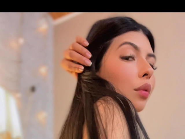 VictoriaGiulias con lunghi capelli neri, una mano tra i capelli, eyeliner, rossetto rosa, sorriso dolce, espressione delicata, pelle chiara, sfondo leggermente sfocato, illuminazione calda, bokeh delicato..