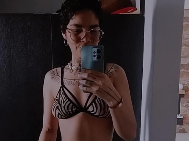 AnaRoa with curly dark hair, glasses, and tattoos, wearing a zebra-patterned bikini top, taking a mirror selfie with a blue phone, showing her small boobs and subtle ass.