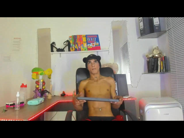 MateusSheenn, shirtless with a black hat, sits in a chair holding a flat screen, looking up, with a slim, toned body and a navel piercing, in a cluttered room.